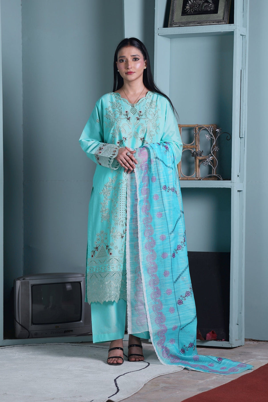 A woman stands indoors wearing a light turquoise traditional outfit with matching pants and a long patterned dupatta. The room has pale blue walls, built-in shelving, a small vintage television, and a rug on the floor.