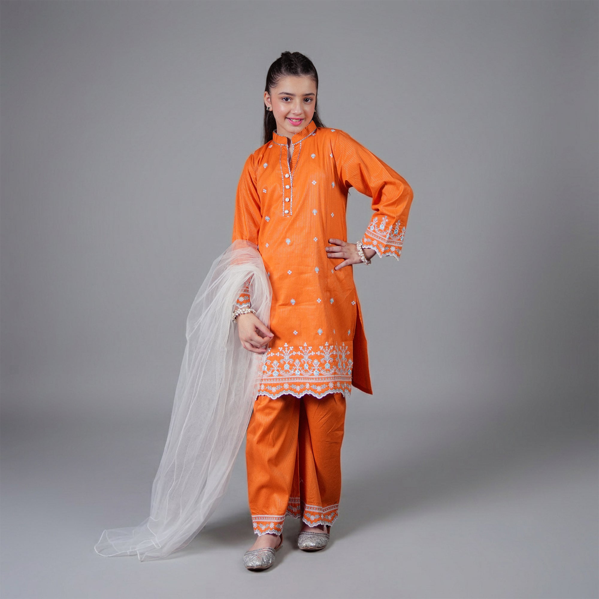 A young girl modeling a traditional bright orange salwar kameez suit with white embroidery, matching orange pants, and a white dupatta, standing against a solid gray studio background.